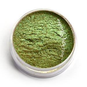 May include: A small, clear jar filled with shimmering, olive-green eyeshadow. The product has a textured surface and appears finely milled. The text "JetAge Studio" is printed on the side of the jar in a cursive font.