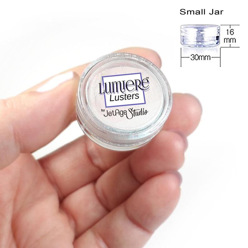 May include: A small jar of Lumiere Lustre by Jet Age Studio. The jar is 30 mm in diameter and 16 mm tall. The jar is filled with a white iridescent powder.