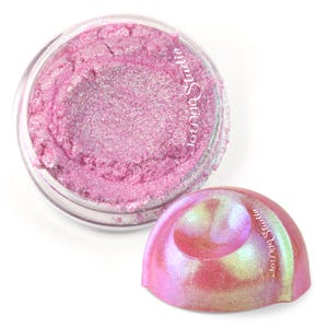 May include: A small, round, clear plastic jar filled with loose, shimmering pink cosmetic powder. The jar has a matching pink, iridescent, dome-shaped lid with the text Jenga Studio printed on it. The powder has a fine, glittery texture.