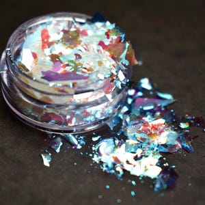 May include: A small, clear plastic jar filled with iridescent, multi-colored flakes. The flakes are a mix of blue, red, and white, with a shimmering, reflective quality. Some flakes have spilled out onto a dark surface.
