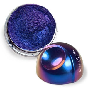 Pode incluir: Um pequeno frasco de pó de pigmento azul e roxo iridescente. O frasco está aberto e o pó é visível. O frasco está sobre uma superfície branca. O frasco é etiquetado com "Joung Studio".