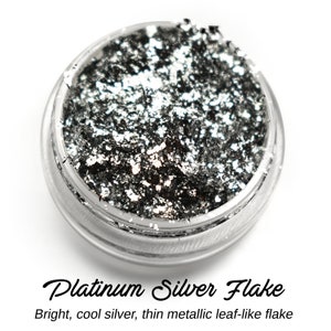 May include: A small jar filled with silver metallic flakes. The flakes are thin and have a cool, bright silver color.