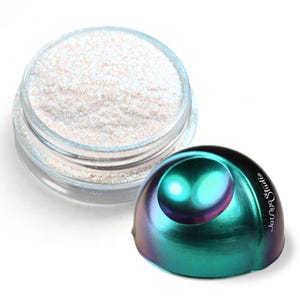 Puede incluir: Un tarro transparente lleno de polvo brillante de color claro se encuentra junto a un objeto iridiscente en forma de cúpula de color verde azulado y púrpura. El objeto tiene un diseño de perla y el texto "Lounge Studio" en su lado.