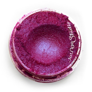 May include: A jar of vibrant, shimmering, deep magenta eyeshadow. The product has a textured, granular appearance with hints of blue iridescence. The jar is clear, and the text "Jetnge Studio" is printed on the side.