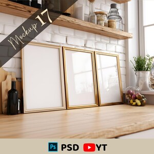 May include: Three gold-framed rectangular picture frames are displayed on a wooden surface. The frames are empty, with a white interior. A wine bottle and cutting board are on the left. The background includes a white brick wall and a wooden shelf.