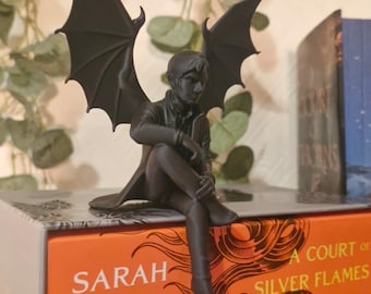 Puede incluir: Una figura gris oscuro de una figura alada, sentada sobre una pila de libros. Los libros se titulan "A Court of Silver Flames", "A Court of Frost and Starlight" y "A Court of Wings and Ruin" de Sarah J. Maas. La figura tiene alas de murciélago.