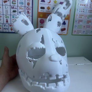 Puede incluir: Una máscara blanca impresa en 3D de un personaje de conejo con una apariencia desgastada y deteriorada. La máscara presenta grandes cuencas oculares huecas, un hocico prominente y dos orejas largas, una de ellas doblada, con bordes irregulares.