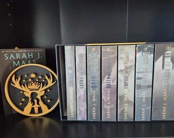 Könnte beinhalten: Eine Sammlung von Fantasy-Büchern von Sarah J. Maas, darunter "Throne of Glass" und "Kingdom of Ash". Die Bücher werden in einer schwarzen Box neben einem Buch und einem gold-schwarzen Zierornament mit Hirschmotiv präsentiert.
