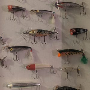 May include: A white framed display case filled with vintage fishing lures. The lures are in various colors, including silver, red, black, and yellow. Each lure has multiple hooks. The case is mounted on a light brown wall.