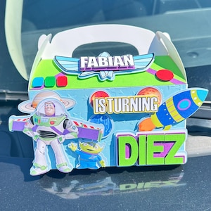 May include: A white cardboard gift box featuring a space-themed design with cutouts of a space ranger and an alien. The box displays the text "FABIAN", "IS TURNING", and "DIEZ" in colorful, layered lettering, along with planets and a rocket ship.