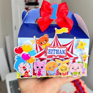 Pode incluir: Uma caixa de lembrancinhas azul e branca com tema de circo, com um laço vermelho e o número 1. A caixa apresenta personagens de desenhos animados e o nome "EITHAN". Balões coloridos e um design de tenda de circo também são visíveis.