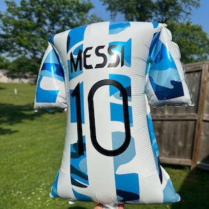 May include: A blue and white foil balloon shaped like a soccer jersey. The jersey has the name "MESSI" and the number "10" printed on it. The balloon is held up by a person's hand in front of a green lawn and blue sky.
