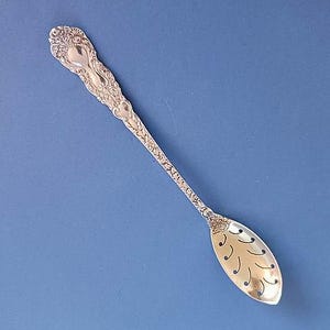 May include: A silver spoon with an ornate handle and a slotted, oval bowl. The handle features detailed floral and scrollwork patterns. The spoon is set against a solid blue background, highlighting its intricate design and craftsmanship.