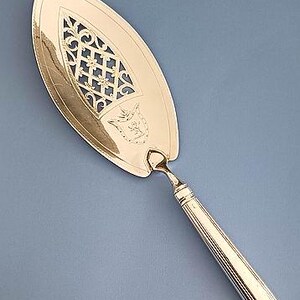 May include: A gold-toned cake server featuring an elongated, pointed blade with an intricate, openwork lattice design and a small engraved crest. The handle is slender, cylindrical, and decorated with vertical fluted lines along its length.