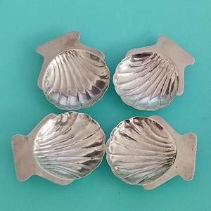 May include: A set of four silver-toned metal dishes shaped like scallop shells. Each dish features a concave center with radiating ridges and a flat, rectangular tab at the top edge. The metallic surface has a reflective, polished finish.
