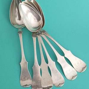 May include: A set of six antique silver spoons with elongated bowls and decorative handles. Each spoon handle is engraved with the initials "EM". The spoons are arranged on a teal background.