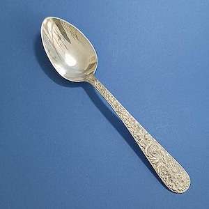 May include: A silver spoon with an oval bowl and a handle decorated with floral patterns. The spoon is set against a solid blue background, highlighting its reflective surface and intricate design.