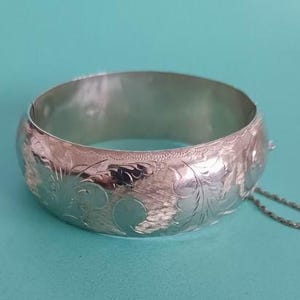 May include: A silver hinged bangle bracelet with an engraved floral design. The bracelet is round and has a clasp with a safety chain. The background is a solid teal color.