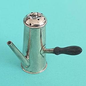 May include: A small, silver-colored coffee pot with a black handle and a spout. The pot has a rounded base and a lid with small holes. The handle is made of dark wood. The pot is set against a turquoise background.