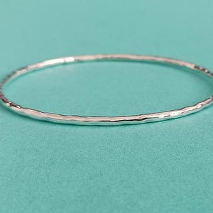 May include: A thin, circular silver bangle bracelet featuring a hammered, textured surface. The metal band has a reflective, polished finish that catches the light, creating subtle variations along the circumference of the continuous, round metal loop.
