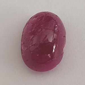 May include: A polished, oval-shaped ruby gemstone. The gem is a deep, rich red color with visible internal inclusions and a glossy surface. The stone is set against a plain, light background.
