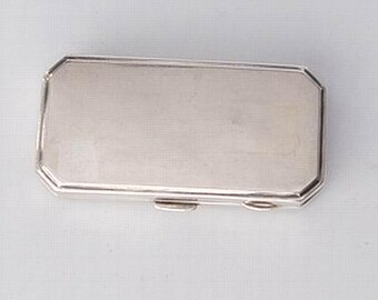May include: A rectangular silver-colored box with a hinged lid. The box has a geometric design with beveled edges. The surface of the box is smooth and reflective, suggesting a polished finish. The box is likely made of metal.