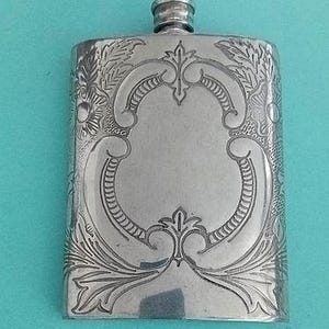May include: A rectangular silver-toned metal flask featuring an intricate engraved pattern of scrolling flourishes and floral motifs. The flask has a smooth central area for personalization and a small, cylindrical screw-top cap at the center top.