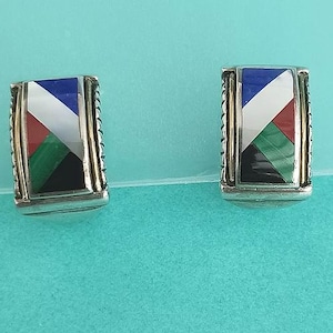 May include: A pair of rectangular earrings with a silver and gold frame. Each earring features a geometric design with blue, white, red, green, and black segments. The earrings are displayed on a turquoise surface.