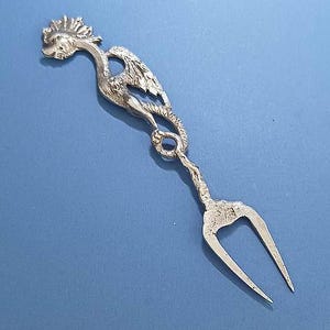 May include: A silver-colored serving fork with a decorative bird design. The fork features a bird-like figure with a detailed head, wings, and a twisted stem leading to the two-pronged fork. The background is a solid blue.