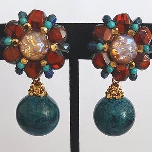 May include: A pair of dangle earrings with a large, multi-colored flower design and a teal-colored ball hanging from each flower. The flowers are made of red, gold, turquoise, and blue beads.