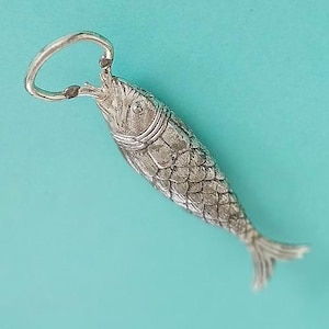 May include: A silver-toned metal bottle opener shaped like a fish. The fish features detailed scales, a defined head, and a tail fin. A circular metal ring is attached to the mouth of the fish, serving as the functional part of the bottle opener.