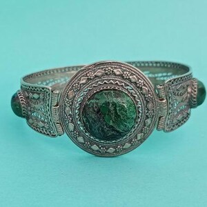 May include: A silver-toned hinged bracelet with intricate filigree and a large, round, green stone center. Two smaller green stones are set on either side of the central stone. The bracelet has a detailed, ornate design.