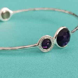 May include: A silver bangle bracelet featuring a hammered texture and three circular settings. The settings contain faceted dark purple stones, with the smaller center stone surrounded by a delicate border of tiny clear pavé crystals.
