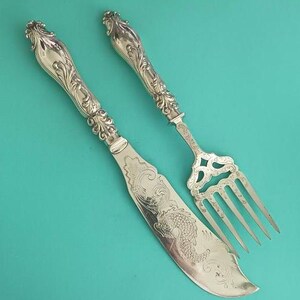 May include: Silver butter knife and fork set with ornate handles and decorative etching. The knife blade features a detailed fish design. The fork has a decorative openwork design. The set is displayed on a teal background.