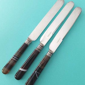May include: Three butter knives with silver blades and dark brown handles with white and clear accents. The handles have a rounded shape and are attached to the blades with a silver metal collar. The knives are arranged diagonally on a turquoise background.