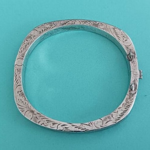 May include: A silver square bangle bracelet with intricate floral engravings. The bracelet has a clasp closure and is displayed against a turquoise background. The metal has a slightly aged appearance, highlighting the detailed craftsmanship.