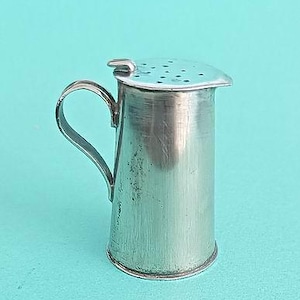 May include: A small, silver-colored creamer with a handle and a perforated lid. The creamer has a cylindrical body and a curved handle. The lid has a small tab for easy opening. The creamer is likely made of metal and has a vintage appearance.
