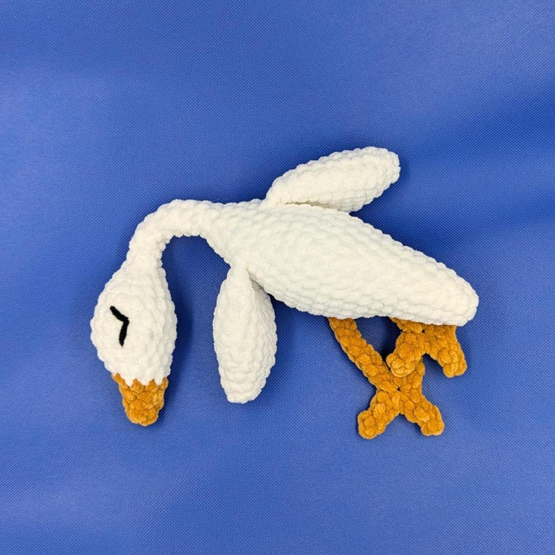 May include: A handmade white crochet swan with a curved neck and a yellow beak. The swan has yellow feet and is lying on a blue surface. The soft, plush toy is a whimsical decorative item.