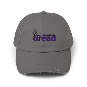 Könnte beinhalten: Eine graue, abgenutzte Baseballkappe mit einem schwarzen und weißen Geistermotiv und dem Text "existential dread" in Lila.