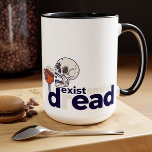 May include: White ceramic mug with a black rim and handle. The mug features a cartoon skull drinking from a coffee cup with the text "existential dread" in navy blue.