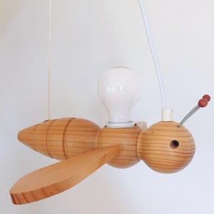 Può includere: Lampada a sospensione a forma di ape in legno. La lampada ha una lampadina al centro e ali ai lati. La testa dell'ape ha un piccolo foro e due antenne rosse. È appesa a un cavo bianco.