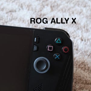 Könnte beinhalten: Schwarzes Handheld-Gaming-Gerät mit dem Text "ROG ALLY X" oben. Das Gerät verfügt über einen schwarzen Bildschirm, ein Steuerkreuz und farbige Aktionstasten: grünes Dreieck, rosa Quadrat, roter Kreis und blaues X.