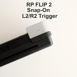 May include: A black and gray Snap-On L2/R2 trigger for a gaming device. The trigger is white with a textured surface and the letters "R2" are visible. The text "RP FLIP 2" is at the top.