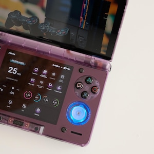 May include: A purple handheld gaming console with a screen displaying a retro game interface. The console features a D-pad, action buttons, and a glowing blue power button. The screen shows 25 FPS and other performance metrics.