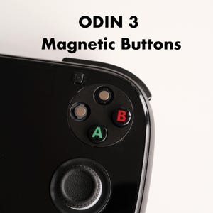 Könnte beinhalten: Nahaufnahme eines schwarzen Handheld-Gaming-Geräts mit dem Text "ODIN 3 Magnetic Buttons". Das Gerät verfügt über ein kreisförmiges Tastenlayout mit A-, B-Tasten in Grün und Rot sowie einen Joystick. Das Gerät befindet sich vor einem weißen Hintergrund.