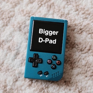 May include: Teal handheld gaming console with a black screen displaying "Bigger D-Pad" in white text. The console features a directional pad, action buttons, and start/select buttons. The device is on a textured, off-white surface.
