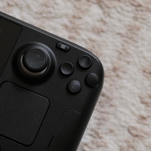 Pode incluir: Um dispositivo de jogo portátil preto com um botão de controle circular, quatro botões e um touchpad. O dispositivo tem uma tela pequena e uma série de pequenos pontos na parte inferior.