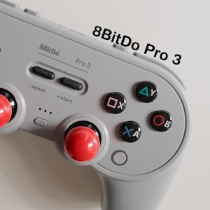 May include: A gray 8BitDo Pro 3 controller with red joysticks and black buttons. The controller has the text "8BitDo Pro 3" printed on it. The buttons are labeled with symbols and letters.