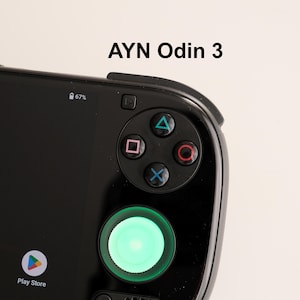 May include: Close-up of a black AYN Odin 3 handheld gaming device. The device features a directional pad, action buttons, and a glowing green joystick. The screen displays a battery percentage and app icons.