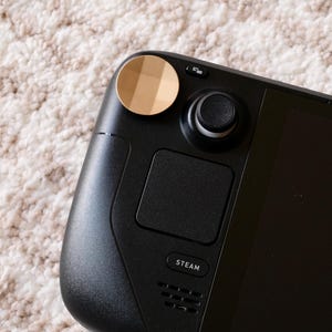 Könnte beinhalten: Nahaufnahme eines schwarzen Handheld-Gaming-Geräts mit goldfarbenem Akzent oben links. Das Gerät verfügt über einen Joystick, ein Touchpad und das Wort "STEAM" unten.